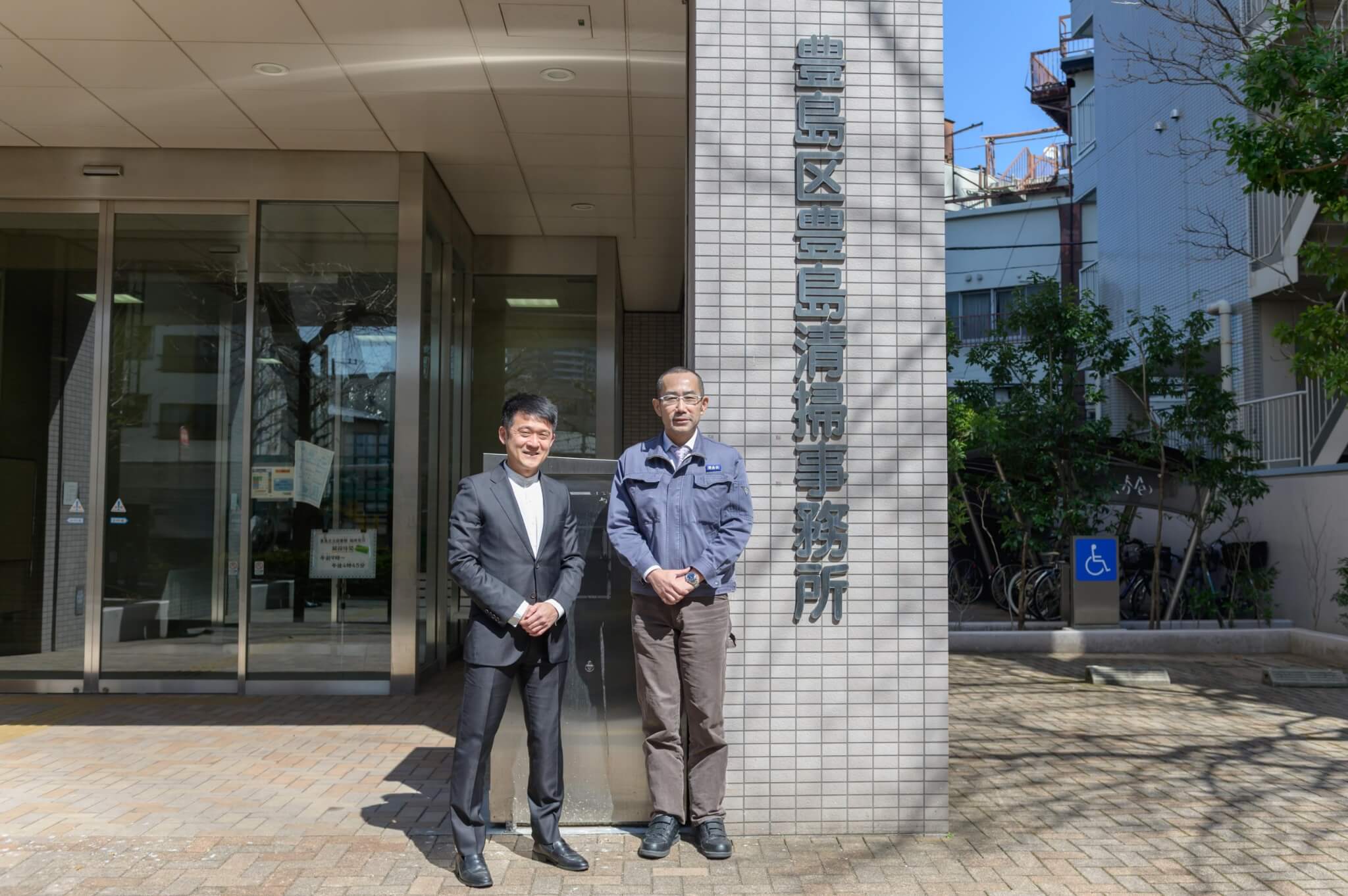 男性が2人が豊島区豊島清掃事務所の前で並んでいる写真